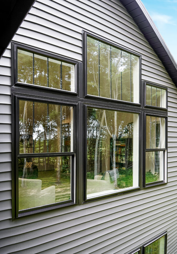 Black double-hung and picture windows on a house, reflecting greenery and outdoor furniture.