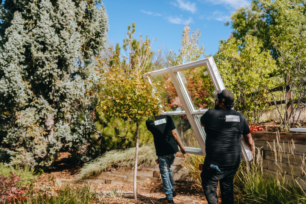 Two Zen Windows installers carry a large window through lots of greenery at a customer's home.